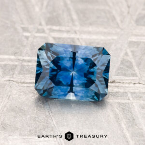 A 1.45-carat heated Montana sapphire, rectangular and blue with intricate facets, is displayed on a textured metallic surface. The text "EARTH'S TREASURY" appears at the bottom.