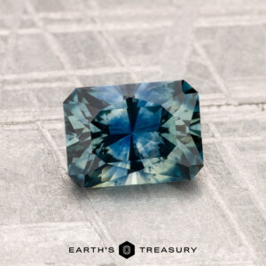 A 1.33-carat heated Montana sapphire, cut in a rectangular facet and showcasing a blue-green hue, is displayed on a textured gray surface with the Earth’s Treasury logo at the bottom.