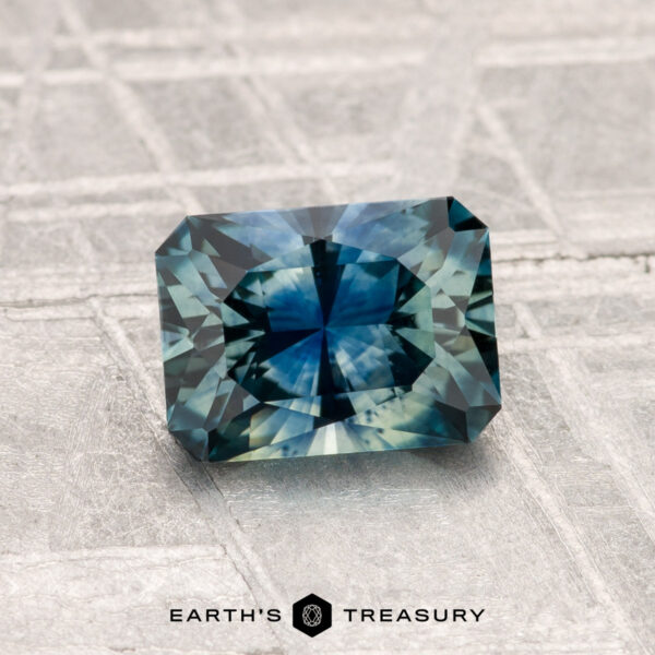 1.33-Carat Montana Sapphire (Heated) A 1.33-carat heated Montana sapphire, cut in a rectangular facet and showcasing a blue-green hue, is displayed on a textured gray surface with the Earth’s Treasury logo at the bottom.