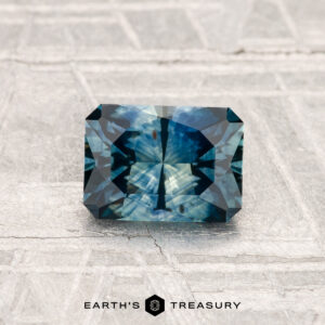 A 1.15-carat, rectangular, faceted Montana sapphire (heated) in blue-green rests on a textured gray surface with "Earth's Treasury" at the bottom.