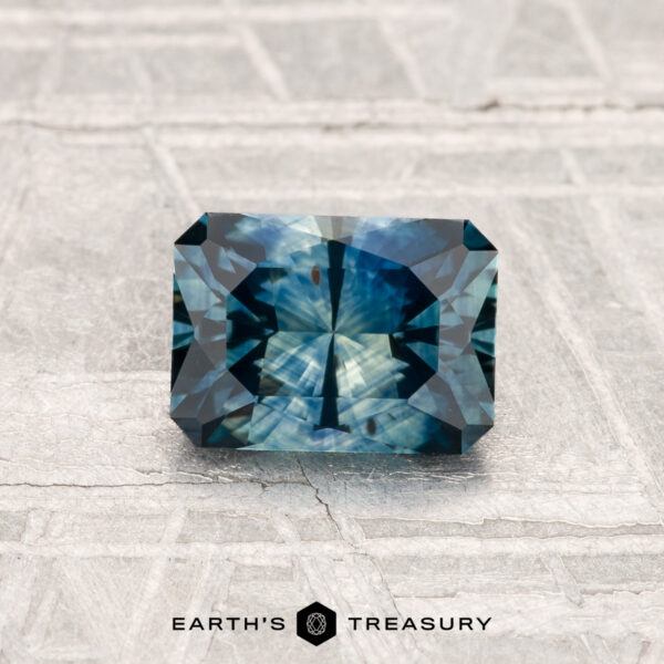 1.15-Carat Montana Sapphire (Heated) A 1.15-carat, rectangular, faceted Montana sapphire (heated) in blue-green rests on a textured gray surface with "Earth's Treasury" at the bottom.