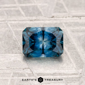 A radiant-cut, rectangular 1.04-carat heated Montana sapphire in deep blue rests on a textured pale gray surface. "EARTH'S TREASURY" is visible at the bottom.
