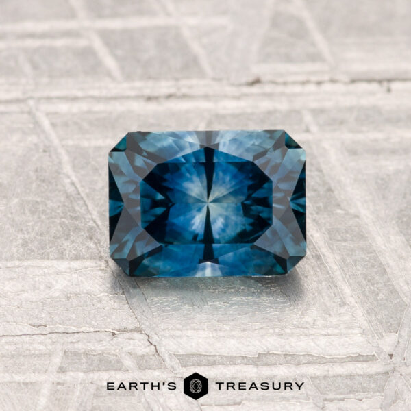 1.04-Carat Montana Sapphire (Heated) A radiant-cut, rectangular 1.04-carat heated Montana sapphire in deep blue rests on a textured pale gray surface. "EARTH'S TREASURY" is visible at the bottom.
