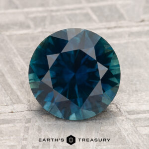 3.04-carat heated Montana sapphire, round-cut and blue-green, displayed on a textured gray surface with "EARTH'S TREASURY" text below.