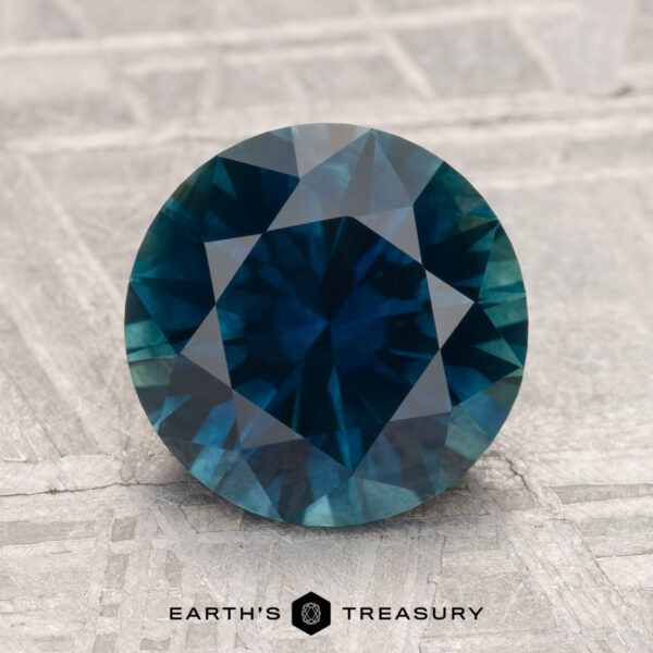 3.04-Carat Montana Sapphire (Heated) 3.04-carat heated Montana sapphire, round-cut and blue-green, displayed on a textured gray surface with "EARTH'S TREASURY" text below.