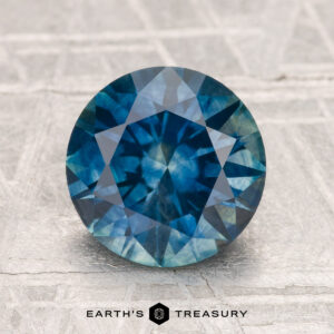 A 2.68-carat heated Montana sapphire, cut in a round brilliant style, displayed on a textured light gray surface with the "Earth's Treasury" logo beneath.