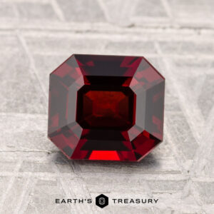 A 1.64-carat, deep red, emerald-cut Spinel gemstone is displayed upright on a textured light gray surface with "EARTH'S TREASURY" written below.