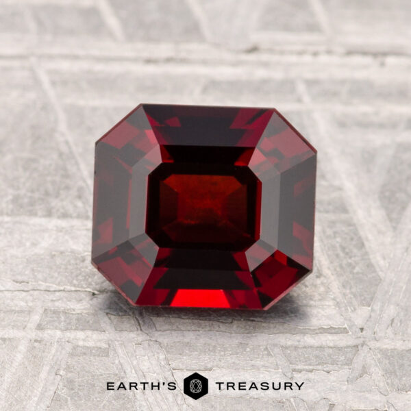 1.64-Carat Spinel A 1.64-carat, deep red, emerald-cut Spinel gemstone is displayed upright on a textured light gray surface with "EARTH'S TREASURY" written below.