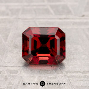 A deep red, emerald-cut 1.33-carat Spinel gemstone is displayed on a textured gray surface with the text "Earth's Treasury" below.