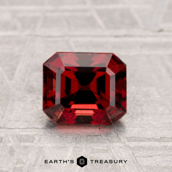 1.33-Carat Spinel A deep red, emerald-cut 1.33-carat Spinel gemstone is displayed on a textured gray surface with the text "Earth's Treasury" below.