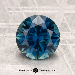 A 2.31-carat heated Montana sapphire with a round brilliant cut is displayed on a light textured surface, featuring the text "EARTH'S TREASURY" at the bottom.