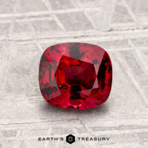 A 1.31-carat cushion-cut Spinel is displayed on a textured, silver-toned surface with "Earth's Treasury" written at the bottom.