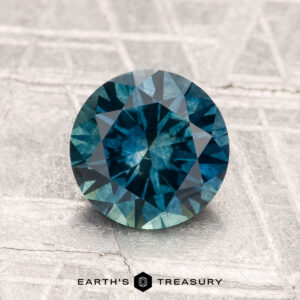 A 1.68-carat heated Montana sapphire, round and blue-green, is shown on a textured light surface with "Earth's Treasury" displayed at the bottom.