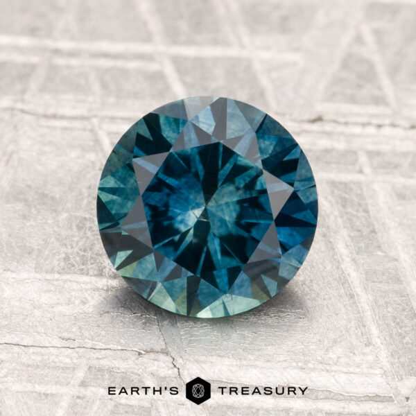 1.68-Carat Montana Sapphire (Heated) A 1.68-carat heated Montana sapphire, round and blue-green, is shown on a textured light surface with "Earth's Treasury" displayed at the bottom.