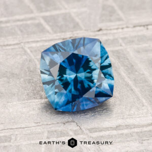 A 1.48-carat heated Montana sapphire in cushion cut with intricate facets is displayed on a textured gray surface, featuring the Earth's Treasury logo at the bottom.