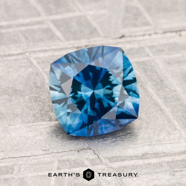 1.48-Carat Montana Sapphire (Heated) A 1.48-carat heated Montana sapphire in cushion cut with intricate facets is displayed on a textured gray surface, featuring the Earth's Treasury logo at the bottom.