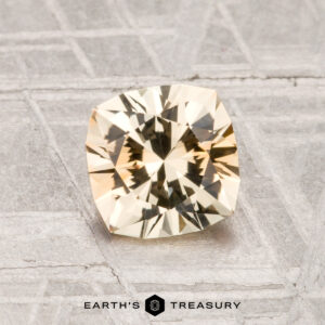 A faceted, light yellow 1.37-carat heated Montana sapphire is displayed on a patterned gray surface with "EARTH'S TREASURY" text at the bottom.