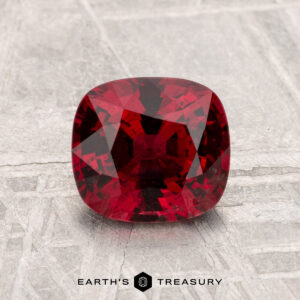 A 1.54-carat cushion-cut red Spinel is displayed on a textured light gray surface with "EARTH'S TREASURY" at the bottom.