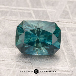 A 2.22-carat heated Montana sapphire with a rectangular, faceted blue-green appearance is displayed on a metallic patterned surface, featuring the text "EARTH'S TREASURY" at the bottom.
