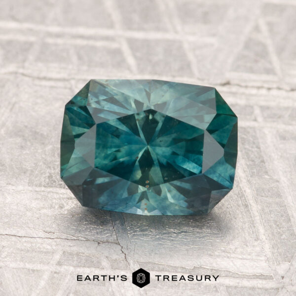 2.22-Carat Montana Sapphire (Heated) A 2.22-carat heated Montana sapphire with a rectangular, faceted blue-green appearance is displayed on a metallic patterned surface, featuring the text "EARTH'S TREASURY" at the bottom.