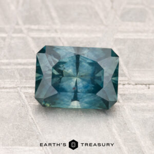 1.52-Carat Montana Sapphire (Heated), a rectangular, faceted blue-green gemstone, displayed on a textured light gray surface with the "Earth's Treasury" logo below.