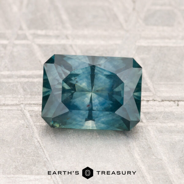 1.52-Carat Montana Sapphire (Heated) 1.52-Carat Montana Sapphire (Heated), a rectangular, faceted blue-green gemstone, displayed on a textured light gray surface with the "Earth's Treasury" logo below.