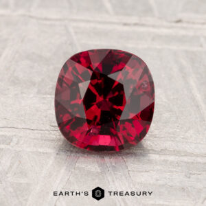 A cushion-cut 1.43-carat Spinel gemstone is displayed on a textured light gray surface with the text "EARTH'S TREASURY" at the bottom.