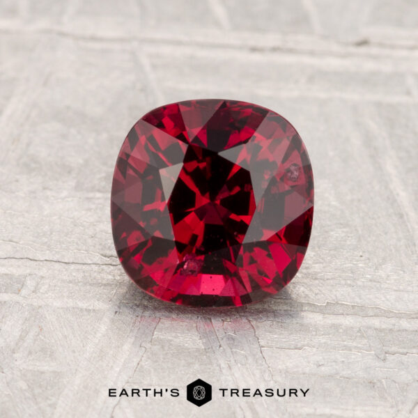 1.43-Carat Spinel A cushion-cut 1.43-carat Spinel gemstone is displayed on a textured light gray surface with the text "EARTH'S TREASURY" at the bottom.