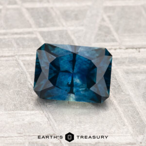 1.33-Carat Montana Sapphire (Heated), a rectangular blue-green gemstone with geometric facets, displayed on a textured silvery surface. "Earth's Treasury" text and logo are visible at the bottom.