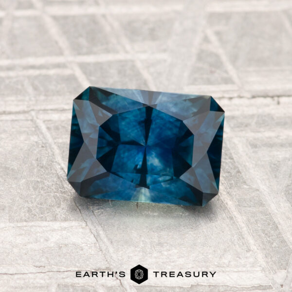 1.33-Carat Montana Sapphire (Heated) 1.33-Carat Montana Sapphire (Heated), a rectangular blue-green gemstone with geometric facets, displayed on a textured silvery surface. "Earth's Treasury" text and logo are visible at the bottom.