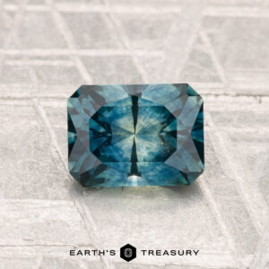 A rectangular, blue-green 1.29-carat heated Montana sapphire is displayed on a textured surface with the "Earth's Treasury" logo at the bottom.