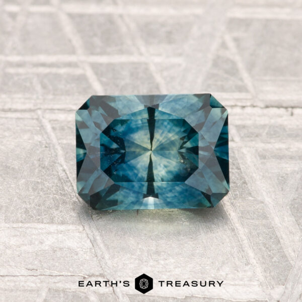 1.29-Carat Montana Sapphire (Heated) A rectangular, blue-green 1.29-carat heated Montana sapphire is displayed on a textured surface with the "Earth's Treasury" logo at the bottom.
