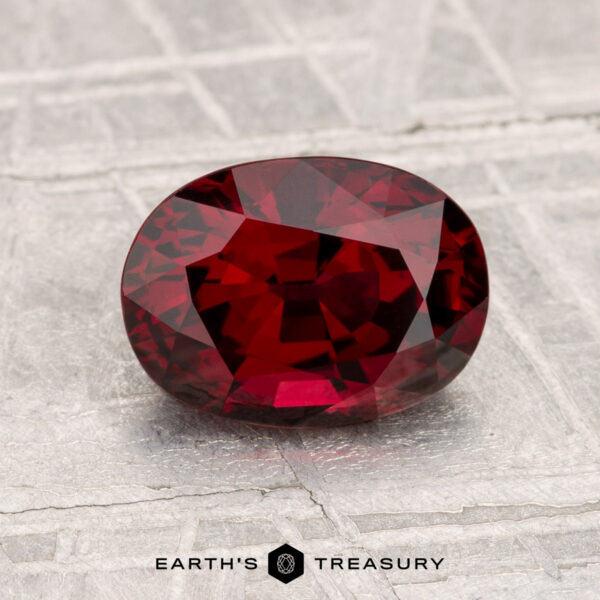 A 1.86-carat, deep red, oval-cut spinel is displayed on a textured gray surface. "Earth's Treasury" is written below the gemstone.