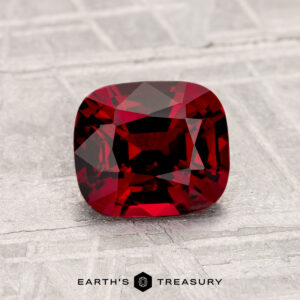 A cushion-cut 1.32-carat Spinel gemstone displayed on a textured light gray surface with the Earth’s Treasury logo at the bottom.