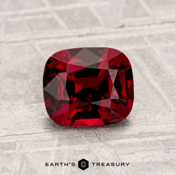 1.32-Carat Spinel A cushion-cut 1.32-carat Spinel gemstone displayed on a textured light gray surface with the Earth’s Treasury logo at the bottom.