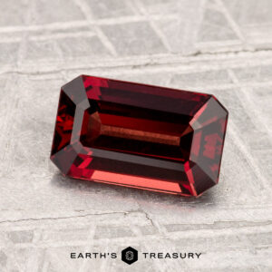 A 1.74-carat spinel, rectangular and deep red with emerald cut facets, rests on a textured gray surface. The "EARTH'S TREASURY" logo appears below.