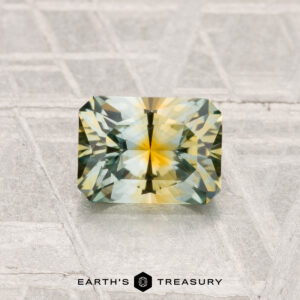 A rectangular, multi-colored 1.16-carat Montana Sapphire (Heated) with yellow and green hues is displayed on a textured surface with "EARTH'S TREASURY" below.