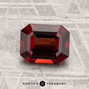 A 1.57-carat, emerald-cut deep red spinel rests on a textured gray surface with "Earth's Treasury" displayed at the bottom.