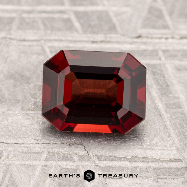 1.57-Carat Spinel A 1.57-carat, emerald-cut deep red spinel rests on a textured gray surface with "Earth's Treasury" displayed at the bottom.