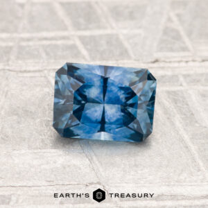 A 1.06-carat heated Montana sapphire, rectangular and intricately faceted, rests on a textured light gray surface. The text "EARTH'S TREASURY" appears at the bottom.