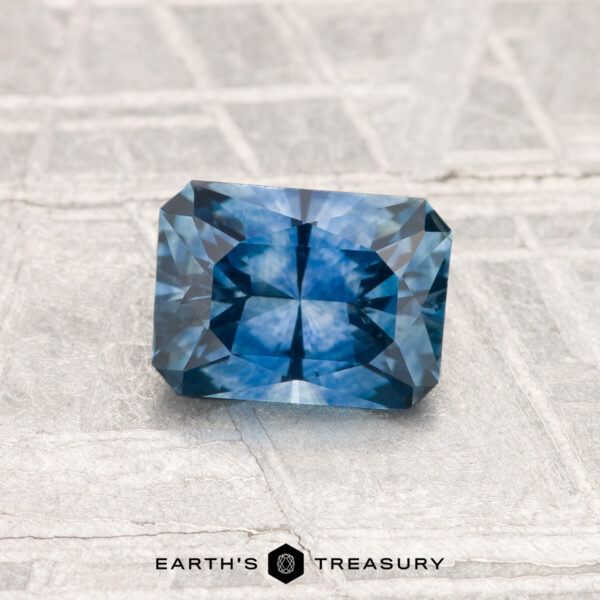 1.06-Carat Montana Sapphire (Heated) A 1.06-carat heated Montana sapphire, rectangular and intricately faceted, rests on a textured light gray surface. The text "EARTH'S TREASURY" appears at the bottom.
