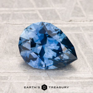 A pear-shaped blue 2.19-carat heated Montana sapphire rests on a textured surface, with the "Earth's Treasury" logo and text at the bottom.