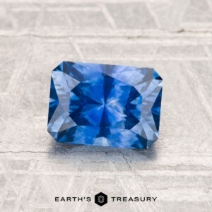 A rectangular 1.57-carat heated Montana Sapphire with step-cut facets is displayed on a textured gray surface, featuring the "Earth's Treasury" logo at the bottom.