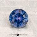 A 1.63-carat heated Montana sapphire with a round brilliant cut is displayed on a textured light gray surface, featuring the Earth’s Treasury logo at the bottom.