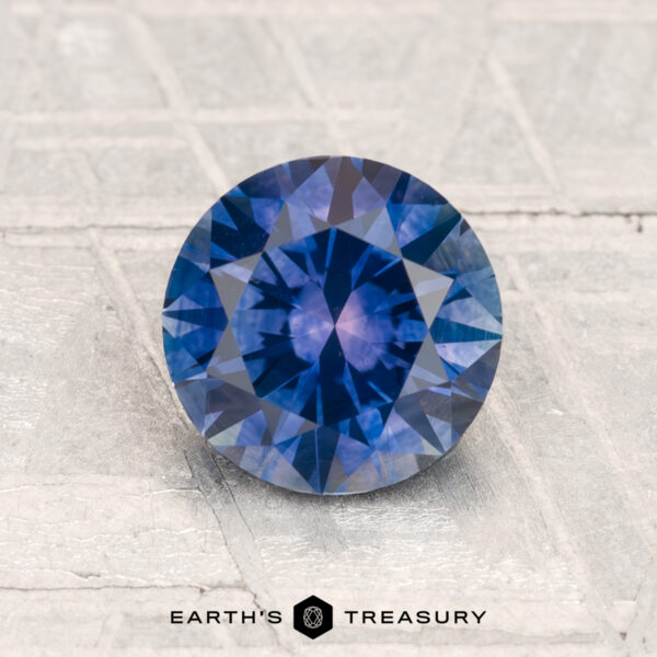 1.63-Carat Montana Sapphire (Heated) A 1.63-carat heated Montana sapphire with a round brilliant cut is displayed on a textured light gray surface, featuring the Earth’s Treasury logo at the bottom.