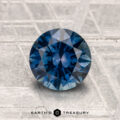 A round, blue 1.63-carat heated Montana sapphire is shown on a textured metallic surface with the "Earth's Treasury" logo at the bottom.