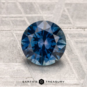 A round, blue 1.63-carat heated Montana sapphire is shown on a textured metallic surface with the "Earth's Treasury" logo at the bottom.