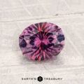 A 1.78-carat heated Montana sapphire, round and faceted with purplish pink hues, rests on a textured light surface. "EARTH'S TREASURY" is displayed at the bottom of the image.