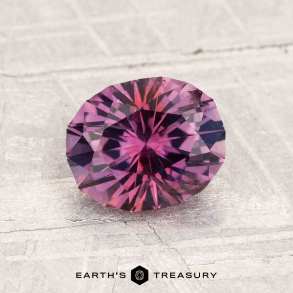 1.78-Carat Montana Sapphire (Heated) A 1.78-carat heated Montana sapphire, round and faceted with purplish pink hues, rests on a textured light surface. "EARTH'S TREASURY" is displayed at the bottom of the image.