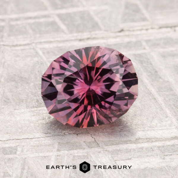 1.78-Carat Montana Sapphire (Heated) A 1.78-carat heated Montana sapphire with a round, faceted cut and pink-purple hue is displayed on a textured light gray surface, featuring the text "EARTH'S TREASURY" at the bottom.